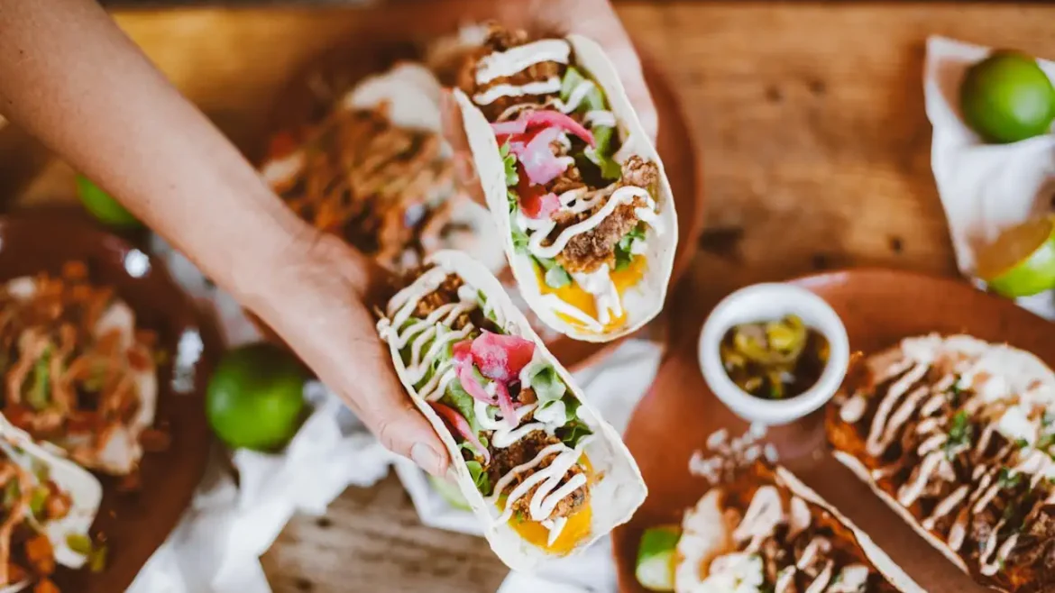 Dłonie trzymają dwa wrapy z warzywami i sosem nad stołem pełnym apetycznych dań.