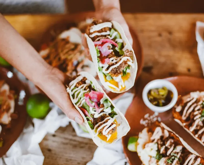 Dłonie trzymają dwa wrapy z warzywami i sosem nad stołem pełnym apetycznych dań.