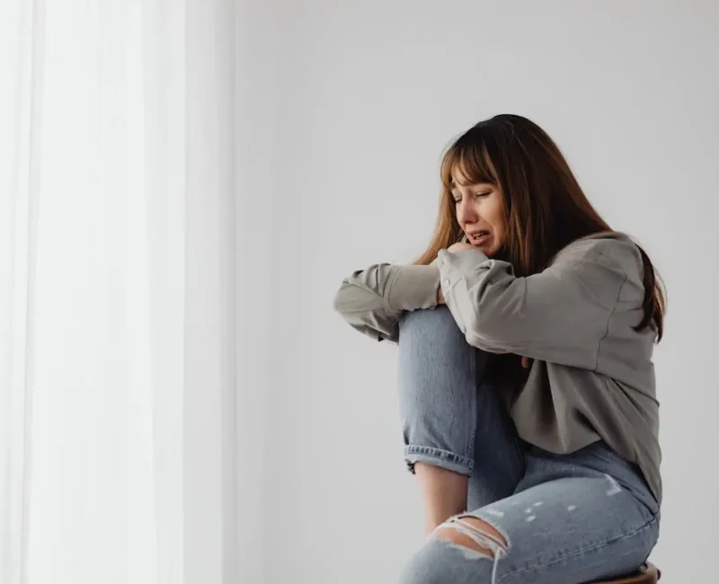 Smutna kobieta siedzi skulona przy jasnej ścianie, opierając głowę na kolanach, co symbolizuje ból emocjonalny i przeżywanie rozstania.