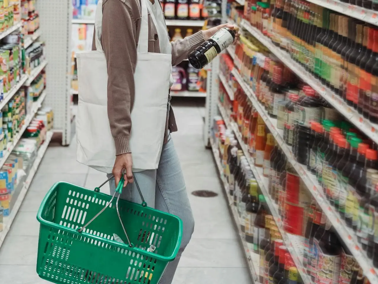 Osoba robi zakupy w supermarkecie, trzyma zielony koszyk i sięga po butelkę z półki z produktami spożywczymi.