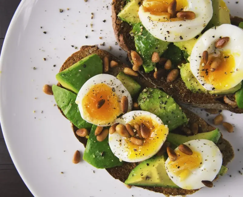 Kanapki z awokado i jajkiem na twardo podane na białym talerzu, posypane ziarnami i pieprzem.